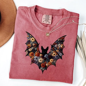 K&ouml;nnte beinhalten: Ein staubrosa Comfort Colors T-Shirt mit einem floralen Fledermaus-Design. Die Fl&uuml;gel der Fledermaus sind mit bunten Blumen gef&uuml;llt. Eine goldene Halskette mit einem kleinen Anh&auml;nger liegt auf dem Shirt. Das Shirt hat ein kleines Etikett am Ausschnitt.