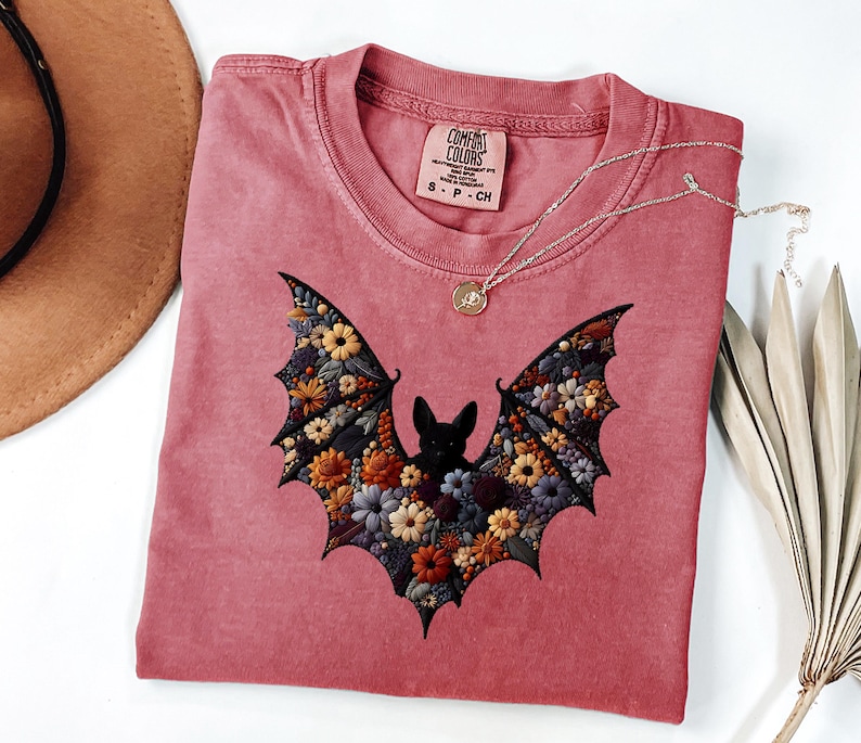 K&ouml;nnte beinhalten: Ein staubrosa Comfort Colors T-Shirt mit einem floralen Fledermaus-Design. Die Fl&uuml;gel der Fledermaus sind mit bunten Blumen gef&uuml;llt. Eine goldene Halskette mit einem kleinen Anh&auml;nger liegt auf dem Shirt. Das Shirt hat ein kleines Etikett am Ausschnitt.