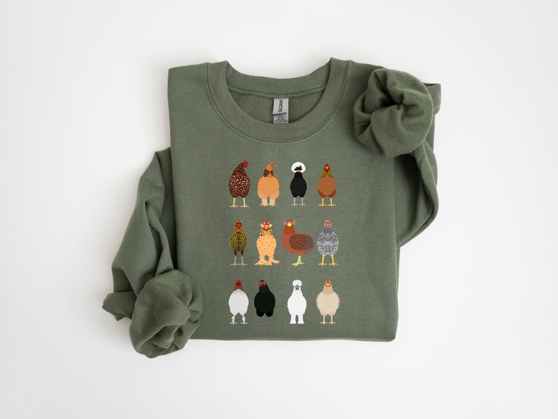 May include: Olive green sweatshirt featuring a grid of twelve illustrated chickens in various colors and patterns. The chickens have different feather designs, including speckled, striped, and solid colors. The sweatshirt has a crew neck and long sleeves.