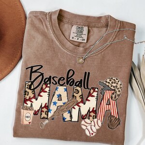 May include: A brown t-shirt with the words "Baseball Mama" in a graphic design. The design includes baseballs, a bat, a helmet, and a glove. A silver necklace with a pendant is draped across the shirt.