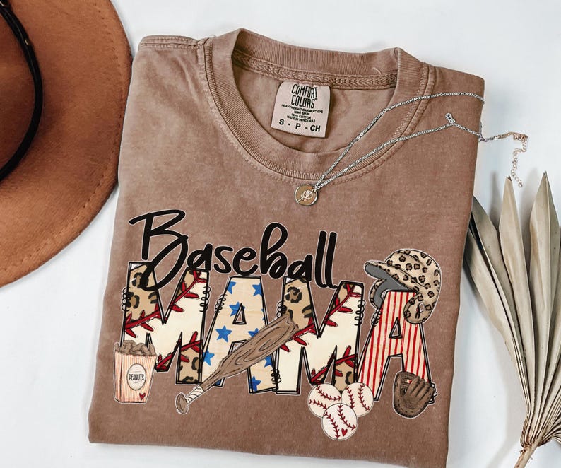 May include: A brown t-shirt with the words "Baseball Mama" in a graphic design. The design includes baseballs, a bat, a helmet, and a glove. A silver necklace with a pendant is draped across the shirt.