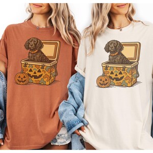 May include: Two t-shirts, one rust-colored and one cream-colored, each featuring a Halloween-themed graphic. The graphic shows a dog in a cooler with a jack-o'-lantern. The cooler has a camouflage pattern with a jack-o'-lantern face.