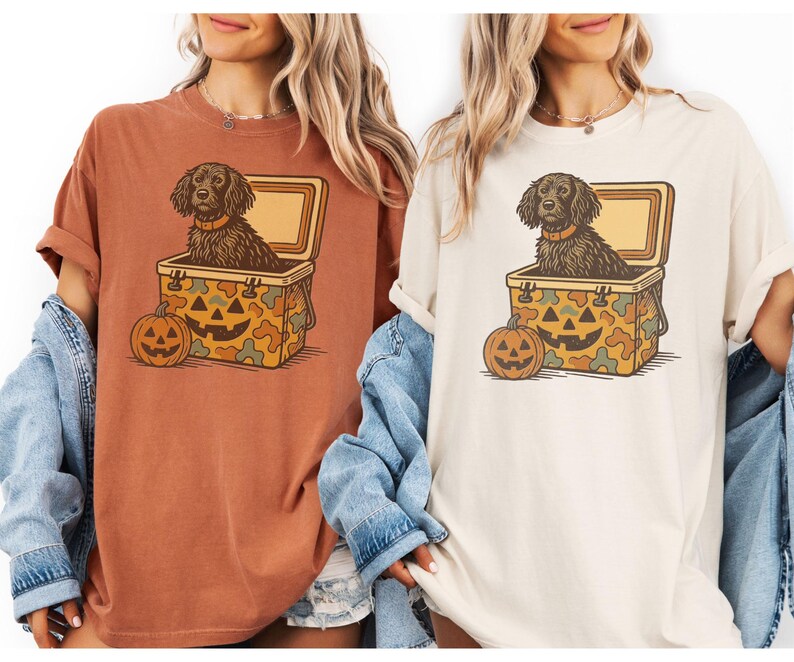 May include: Two t-shirts, one rust-colored and one cream-colored, each featuring a Halloween-themed graphic. The graphic shows a dog in a cooler with a jack-o'-lantern. The cooler has a camouflage pattern with a jack-o'-lantern face.