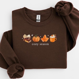 Pode incluir: Camisola castanha escura com um gráfico temático de outono. O gráfico inclui uma pilha de livros, uma abóbora, uma folha de bordo e uma caneca de café. As palavras "cozy season" são impressas abaixo.