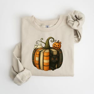 May include: Beige sweatshirt featuring a Halloween-themed graphic. The design includes a pumpkin made of books, a black cat, two ghosts, and a jack-o'-lantern. The sweatshirt has long sleeves and a crew neck.