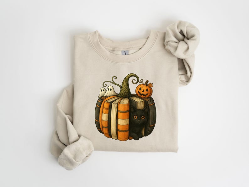 May include: Beige sweatshirt featuring a Halloween-themed graphic. The design includes a pumpkin made of books, a black cat, two ghosts, and a jack-o'-lantern. The sweatshirt has long sleeves and a crew neck.