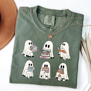 May include: Olive green t-shirt featuring a design of cartoon ghosts reading books. The ghosts are white with black eyes and are holding stacks of books in various colors. A gold necklace with a pendant is draped across the shirt.