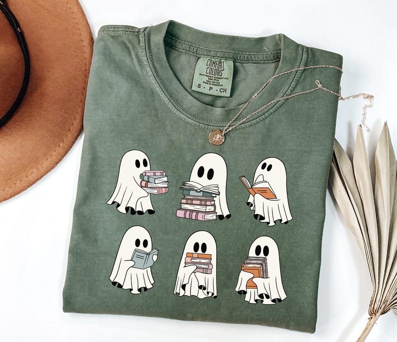 May include: Olive green t-shirt featuring a design of cartoon ghosts reading books. The ghosts are white with black eyes and are holding stacks of books in various colors. A gold necklace with a pendant is draped across the shirt.
