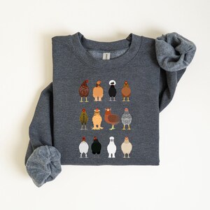 May include: Dark gray sweatshirt featuring a colorful embroidered design of various chicken breeds. The chickens are depicted in a row, each with unique feather patterns and colors. The sweatshirt has long sleeves and a crew neck.