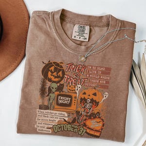 May include: A brown Comfort Colors t-shirt with a Halloween-themed graphic. The graphic includes pumpkins, ghosts, and the text "Trick or Treat" and "Fright Night". A silver necklace with a pendant is draped across the shirt.