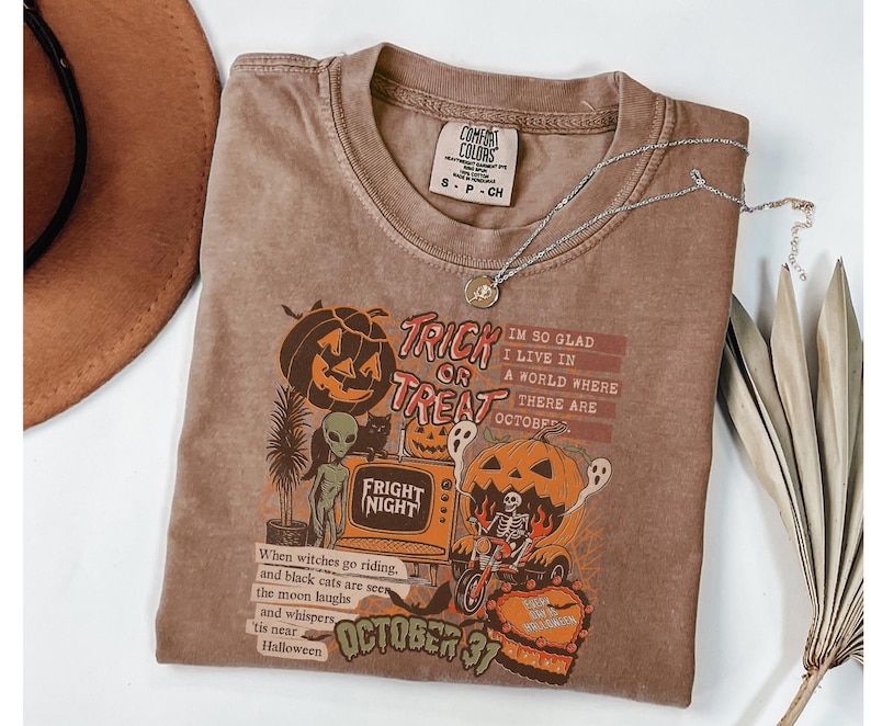 May include: A brown Comfort Colors t-shirt with a Halloween-themed graphic. The graphic includes pumpkins, ghosts, and the text "Trick or Treat" and "Fright Night". A silver necklace with a pendant is draped across the shirt.