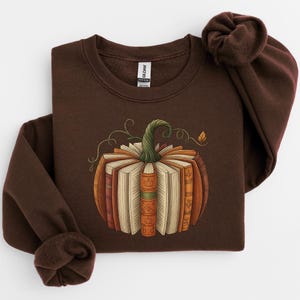 May include: A brown sweatshirt featuring a pumpkin design made of books. The pumpkin has a green stem and is surrounded by swirling vines and a small leaf. The books are various shades of orange, brown, and beige.