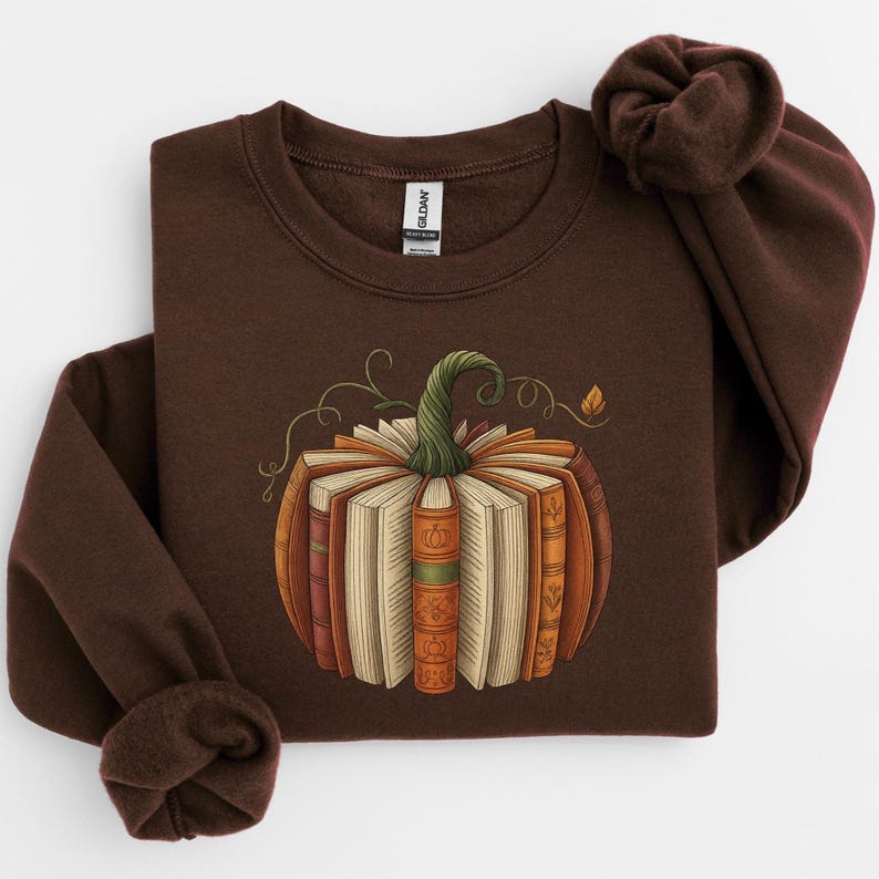 May include: A brown sweatshirt featuring a pumpkin design made of books. The pumpkin has a green stem and is surrounded by swirling vines and a small leaf. The books are various shades of orange, brown, and beige.