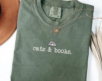 May include: An olive green t-shirt with the words "cats & books" embroidered in white, along with a small graphic of a cat on a book. A gold necklace with a pendant is draped across the shirt. The shirt is folded and placed on a white surface.
