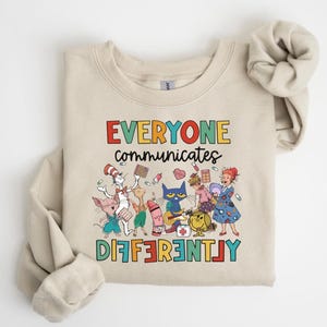 May include: A beige sweatshirt with a colorful graphic featuring characters from popular children's books. The text reads "EVERYONE communicates DIFFERENTLY".