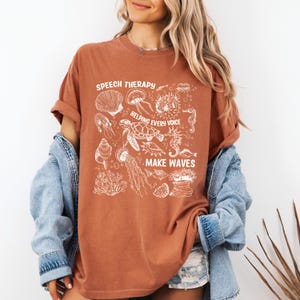 May include: A rust-colored t-shirt with a white graphic featuring various sea creatures and the phrases "SPEECH THERAPY," "HELPING EVERY VOICE," and "MAKE WAVES." The design includes illustrations of seashells, jellyfish, and a sea turtle.