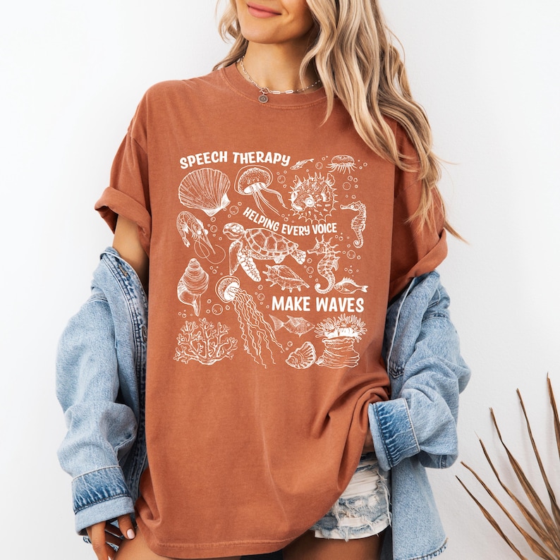 May include: A rust-colored t-shirt with a white graphic featuring various sea creatures and the phrases "SPEECH THERAPY," "HELPING EVERY VOICE," and "MAKE WAVES." The design includes illustrations of seashells, jellyfish, and a sea turtle.