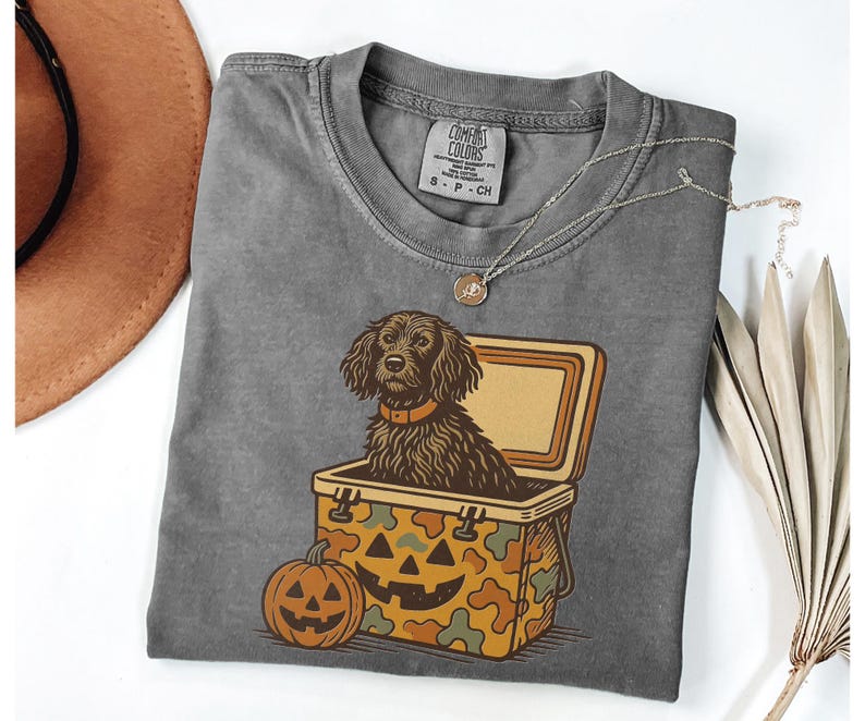 May include: Gray Comfort Colors t-shirt featuring a brown dog inside a jack-o'-lantern cooler. A carved pumpkin sits below the cooler. The shirt has a crew neck and a small tag on the collar.