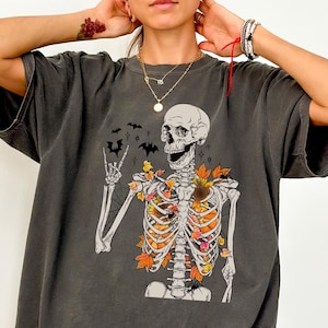 May include: Dark gray t-shirt featuring a skeleton graphic with autumn leaves in the rib cage and bats. The skeleton is making a rock and roll hand gesture. A fun, spooky design for Halloween.