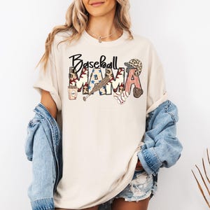May include: Cream-colored t-shirt with the words "Baseball Mama" in a decorative font. The design includes baseball-themed graphics such as a bat, ball, glove, and a baseball cap. The shirt is paired with a denim jacket.