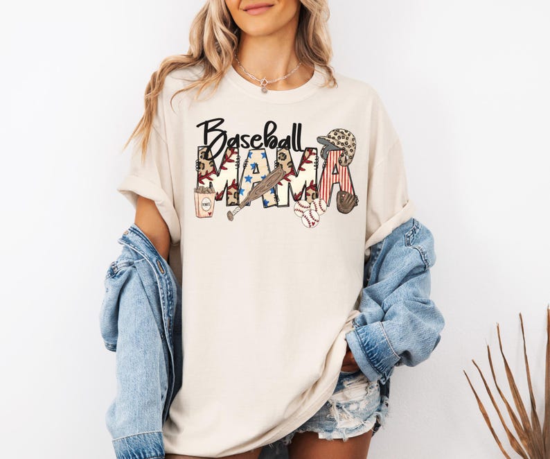 May include: Cream-colored t-shirt with the words "Baseball Mama" in a decorative font. The design includes baseball-themed graphics such as a bat, ball, glove, and a baseball cap. The shirt is paired with a denim jacket.