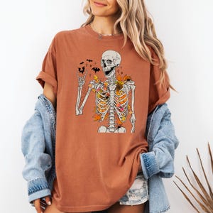 May include: Rust-colored t-shirt featuring a skeleton graphic with autumn leaves and bats. The skeleton is giving a rock-and-roll hand gesture. The design incorporates fall colors and is perfect for Halloween or autumn.