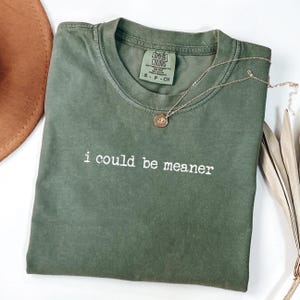 May include: An olive green t-shirt with the phrase "i could be meaner" in white text. A gold necklace with a pendant rests on the shirt. The shirt is folded and has a tag from Comfort Colors.