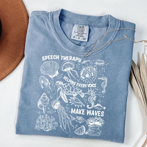 May include: A blue Comfort Colors t-shirt with white graphic illustrations of sea creatures and the text "SPEECH THERAPY HELPING EVERY VOICE MAKE WAVES". A gold necklace and a brown hat are also in the image.