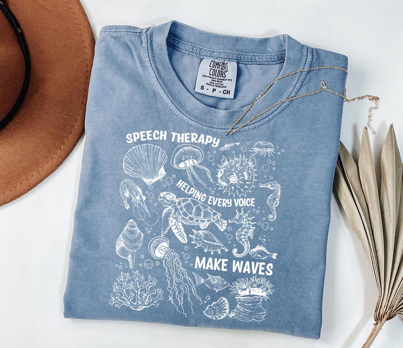 May include: A blue Comfort Colors t-shirt with white graphic illustrations of sea creatures and the text "SPEECH THERAPY HELPING EVERY VOICE MAKE WAVES". A gold necklace and a brown hat are also in the image.