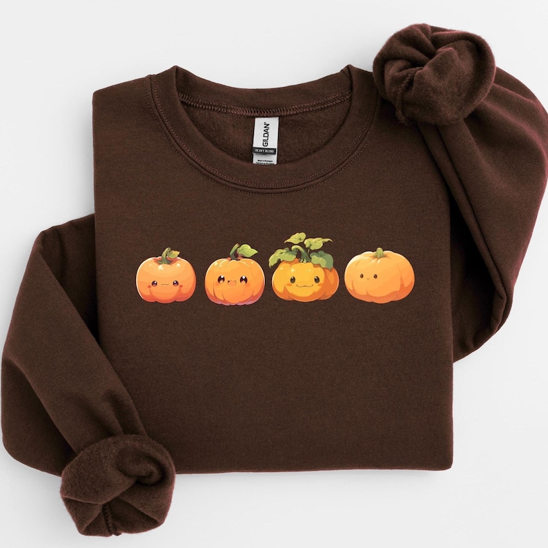Puede incluir: Una sudadera marr&oacute;n oscuro con un gr&aacute;fico de cuatro calabazas de dibujos animados en tonos naranja. Las calabazas tienen caras sonrientes y hojas verdes. La sudadera tiene cuello redondo y mangas largas.