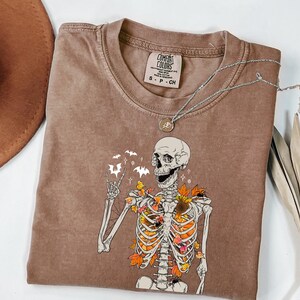May include: Brown Comfort Colors t-shirt featuring a skeleton graphic with autumn leaves and bats. The skeleton is giving a peace sign. A silver necklace with a pendant is on the shirt. A brown hat is in the background.