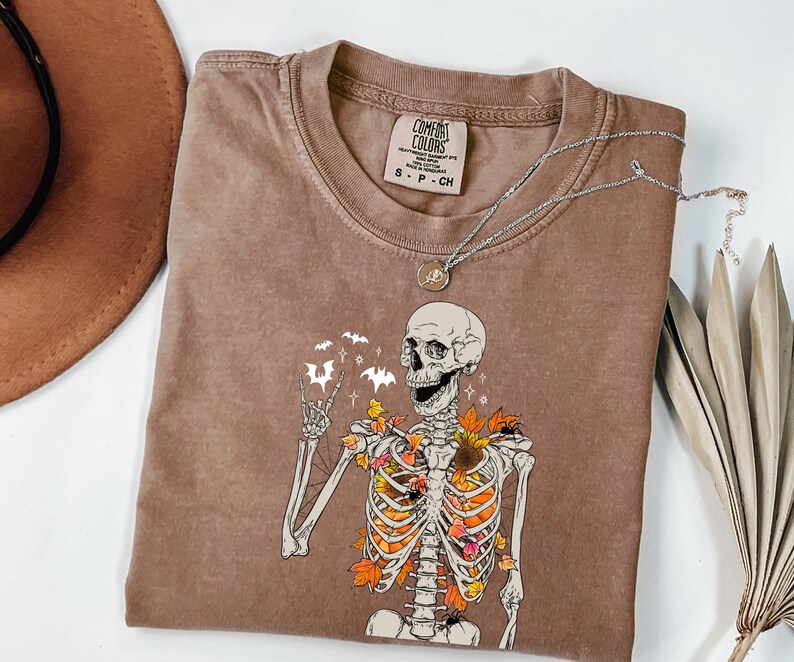 May include: Brown Comfort Colors t-shirt featuring a skeleton graphic with autumn leaves and bats. The skeleton is giving a peace sign. A silver necklace with a pendant is on the shirt. A brown hat is in the background.