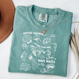 May include: A seafoam green t-shirt with the words "SPEECH THERAPY" and "HELPING EVERY VOICE MAKE WAVES" in white. The shirt features a graphic of various sea creatures. A silver necklace with a pendant is on top of the shirt.