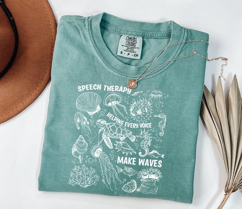 May include: A seafoam green t-shirt with the words "SPEECH THERAPY" and "HELPING EVERY VOICE MAKE WAVES" in white. The shirt features a graphic of various sea creatures. A silver necklace with a pendant is on top of the shirt.