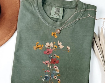 May include: An olive green Comfort Colors t-shirt with a floral cross design. The cross is made of colorful flowers in shades of pink, yellow, and white. A silver necklace with a pendant rests on the shirt.