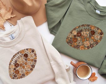 May include: Two crewneck sweatshirts, one cream and one olive green, each featuring a football design made of flowers in shades of brown, tan, and cream. A brown hat and a cup of coffee are also visible.