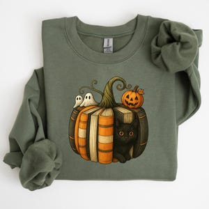 May include: Olive green sweatshirt featuring a Halloween-themed graphic. The design includes a pumpkin made of books, with a black cat peeking out, two ghosts, and a carved pumpkin. The sweatshirt is a comfortable, casual style.