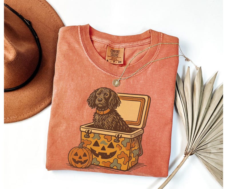 May include: Coral-colored t-shirt featuring a dog illustration inside a cooler with a jack-o'-lantern design. The dog has a brown collar. A brown hat and a gold necklace are also visible.