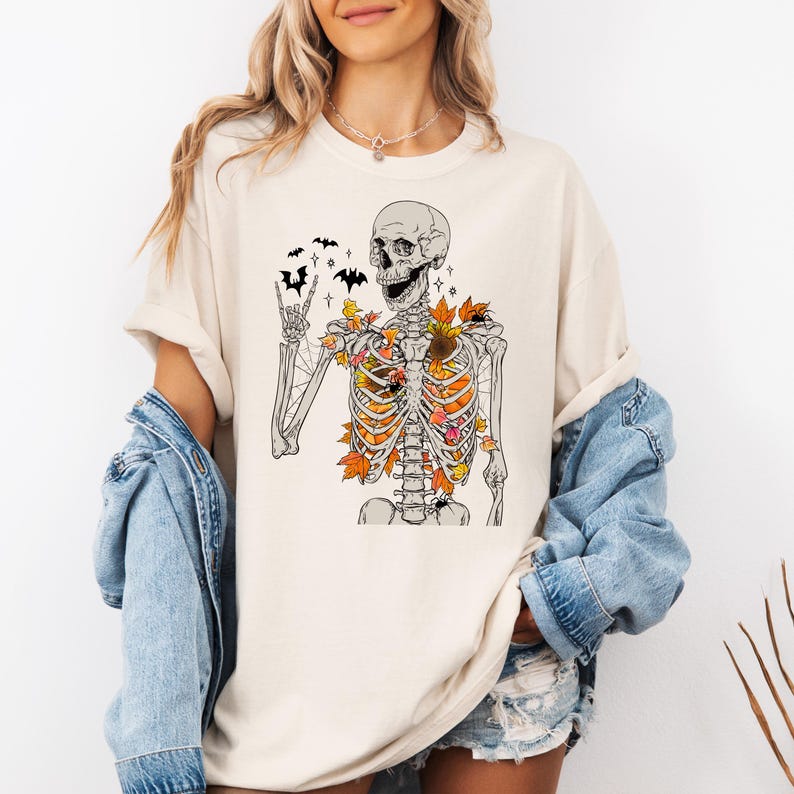 May include: Cream-colored t-shirt featuring a skeleton graphic with autumn leaves in the ribcage and bats above. The skeleton is making a rock-and-roll hand gesture. A spider is also visible. The shirt is a casual, seasonal design.
