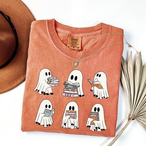 May include: Coral-colored Comfort Colors t-shirt featuring a graphic of six cartoon ghosts, each holding or reading books. The ghosts are white with black eyes and are surrounded by books in various colors. A gold necklace and a brown hat are also visible.