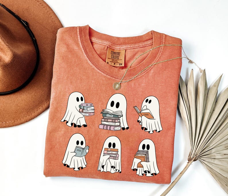 May include: Coral-colored Comfort Colors t-shirt featuring a graphic of six cartoon ghosts, each holding or reading books. The ghosts are white with black eyes and are surrounded by books in various colors. A gold necklace and a brown hat are also visible.
