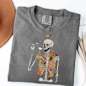 May include: A gray Comfort Colors t-shirt featuring a skeleton graphic with fall leaves and bats. The skeleton is giving a peace sign. A silver necklace with a pendant is on the shirt.