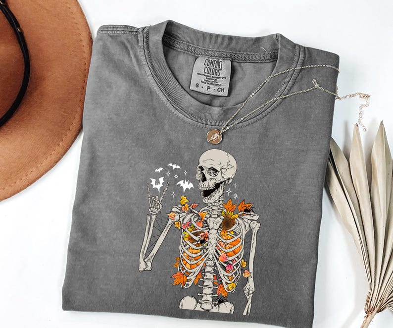 May include: A gray Comfort Colors t-shirt featuring a skeleton graphic with fall leaves and bats. The skeleton is giving a peace sign. A silver necklace with a pendant is on the shirt.