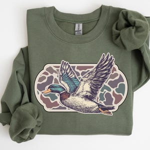 May include: A green sweatshirt with a camouflage pattern and a vintage illustration of a duck in flight.