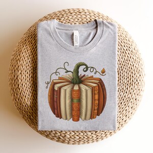 May include: A light gray t-shirt featuring a pumpkin design made of books. The pumpkin has a green stem and is composed of various books with orange, brown, and cream-colored spines. The shirt is laid on a woven, beige surface.