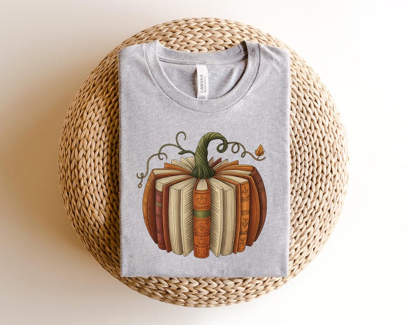 May include: A light gray t-shirt featuring a pumpkin design made of books. The pumpkin has a green stem and is composed of various books with orange, brown, and cream-colored spines. The shirt is laid on a woven, beige surface.