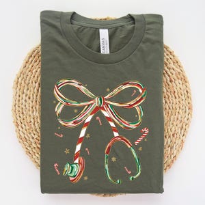Op de afbeelding: Olijfgroen t-shirt met een kerstthema. De afbeelding bevat een strik en een stethoscoop, beide versierd met rood, wit en groen snoeprietstrepen. Kleine snoeprietjes en gouden sterren zijn verspreid over het ontwerp.