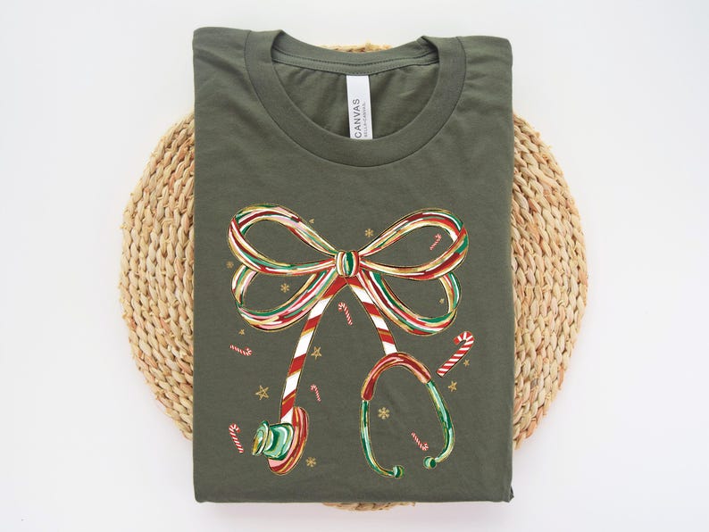 Op de afbeelding: Olijfgroen t-shirt met een kerstthema. De afbeelding bevat een strik en een stethoscoop, beide versierd met rood, wit en groen snoeprietstrepen. Kleine snoeprietjes en gouden sterren zijn verspreid over het ontwerp.