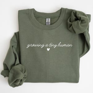 Op de afbeelding: Een olijfgroene sweatshirt met de tekst "growing a tiny human" in witte letters en een klein wit hartje. De top met lange mouwen is opgevouwen, met één mouw opgerold.