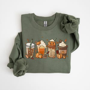 May include: An olive green sweatshirt with a graphic of four autumn-themed drinks: a coffee drink with coffee beans, a latte with whipped cream, a pumpkin spice latte, and a coffee with whipped cream. Pumpkins and autumn leaves are also in the design.